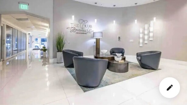 Un área de recepción moderna con una pared curva, un letrero que dice "The Club at Brickell Bay" y un conjunto de asientos con sillas y una mesa.