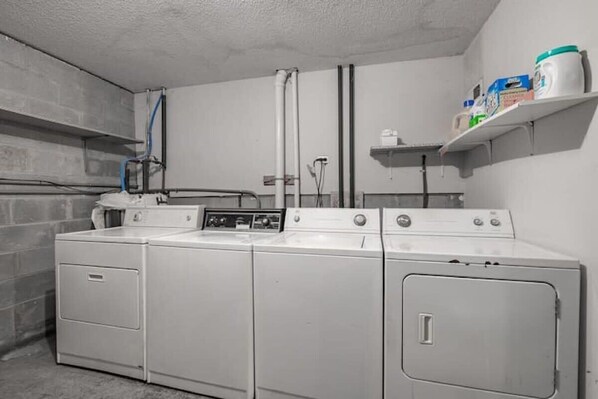 A laundry area with four stacked washers and dryers, a water heater, and shelves with laundry supplies.