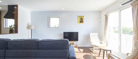 Un salon moderne avec un grand canapé gris, un téléviseur à écran plat et une table basse en bois. On y trouve deux fauteuils et une petite table, et une fenêtre laisse entrer la lumière naturelle.