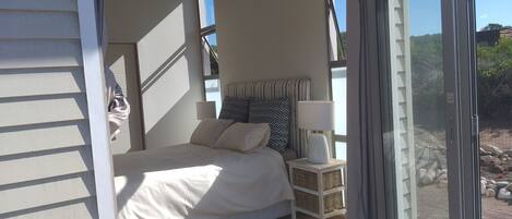 Un dormitorio con cama, almohadas, una mesita de noche, lámpara y un ventanal corredizo que da a un espacio exterior con rocas.