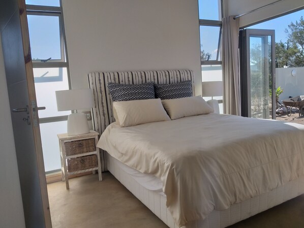 A bedroom with a bed, bedside lamps, a nightstand, and a view through a large window. - Forever Ocean - A Dreamer’s Paradise (Kleinkrantz)
