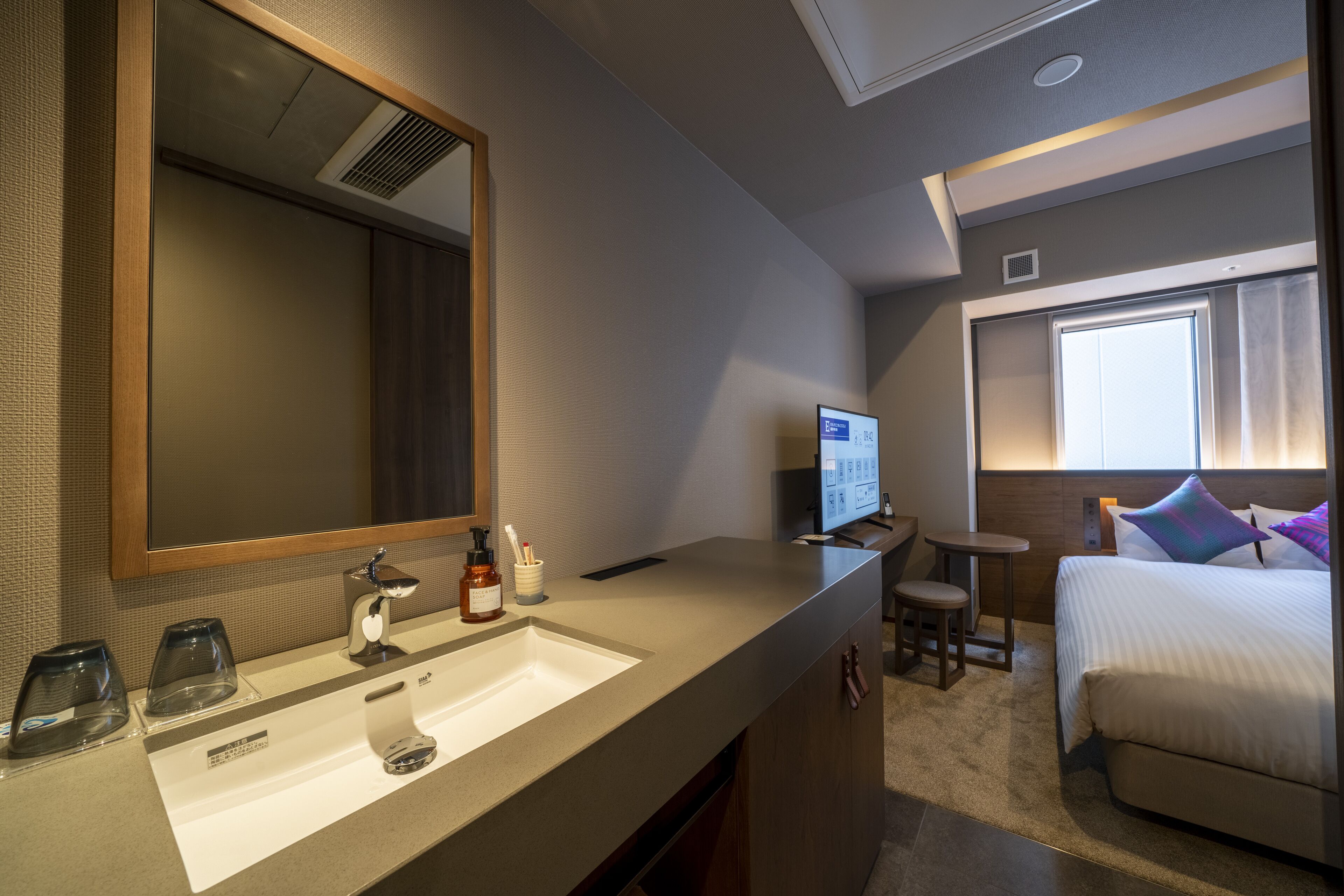 A hotel room with a large mirror, a sink, a small table, a bed, and a window with curtains.