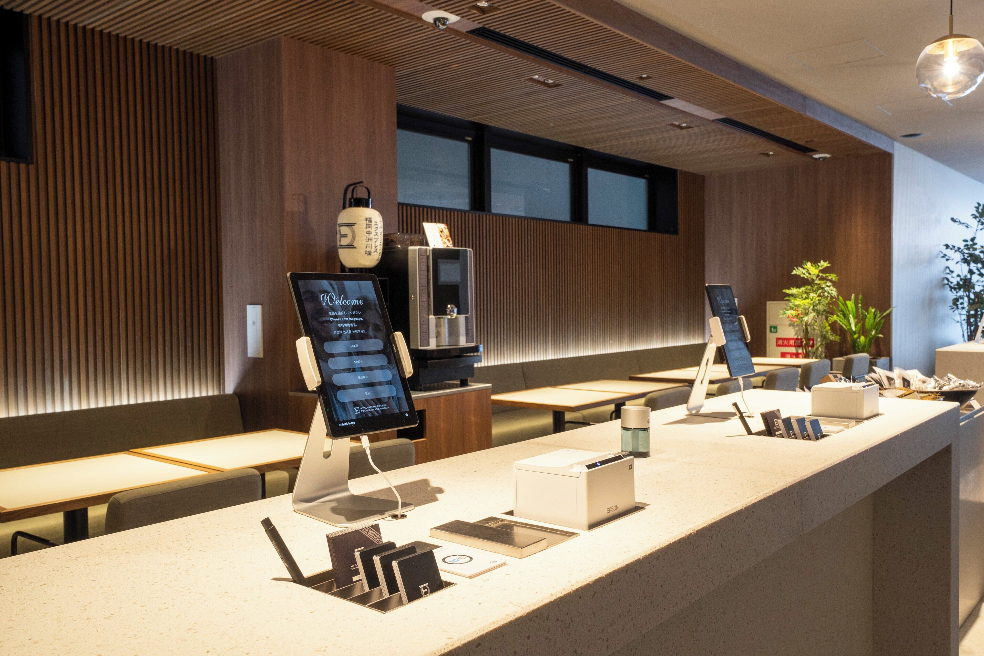 A modern reception area with a long counter, digital check-in kiosks, seating, and a potted plant.