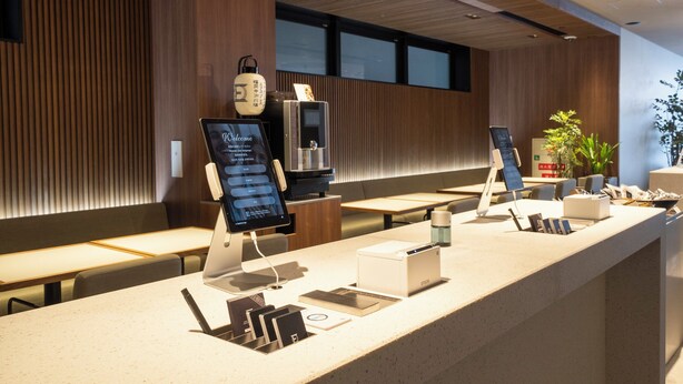 A modern reception area with a long counter, digital check-in kiosks, seating, and a potted plant.