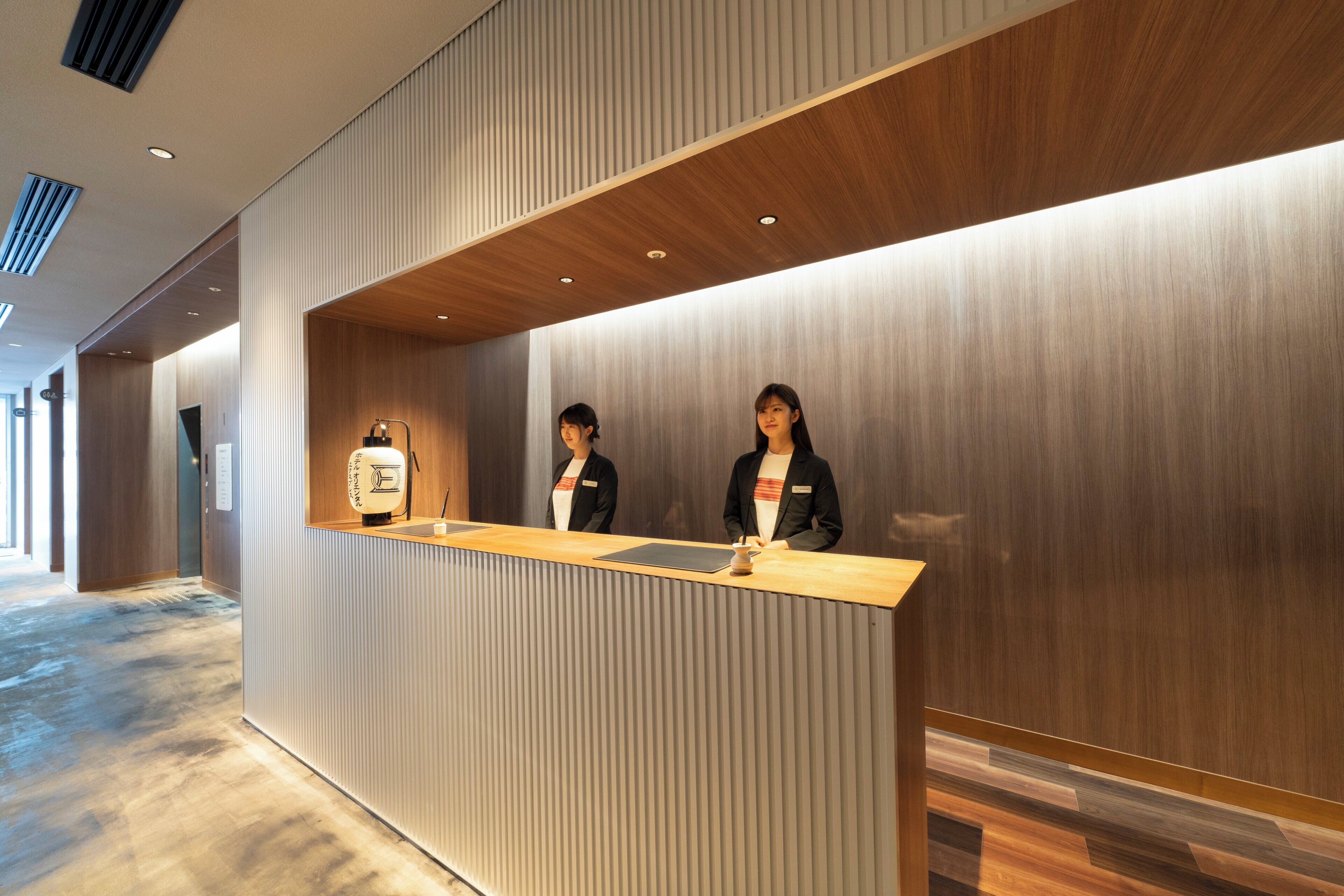 A reception desk with two staff members, a wooden counter, and a modern interior with vertical paneling and recessed lighting.