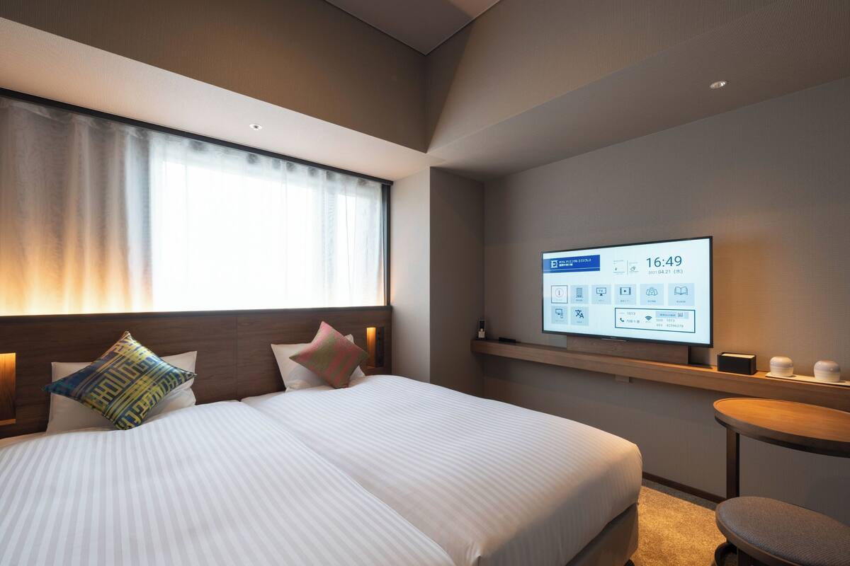 A modern hotel room with a large bed, a bedside table with a lamp, a TV mounted on the wall displaying the time, and a small round table with a chair.