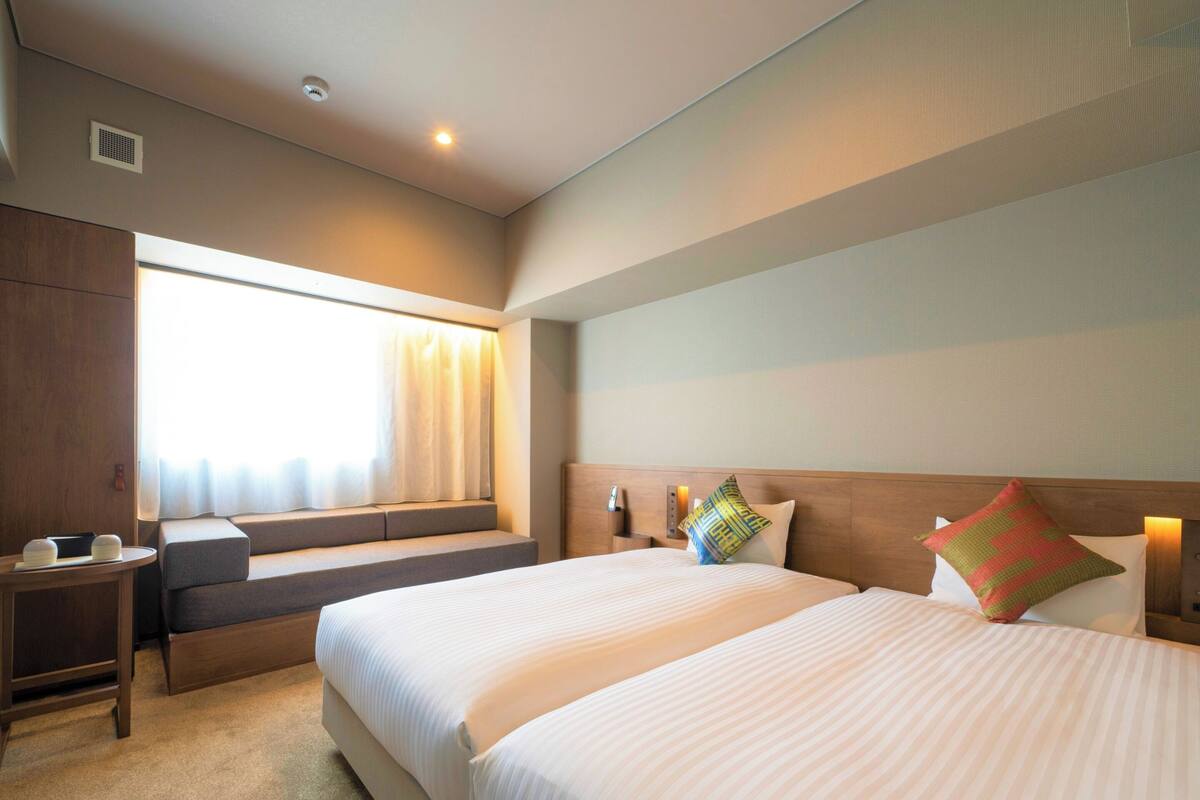 A hotel room with two beds, a window with curtains, a bedside table, and a headboard with decorative pillows.