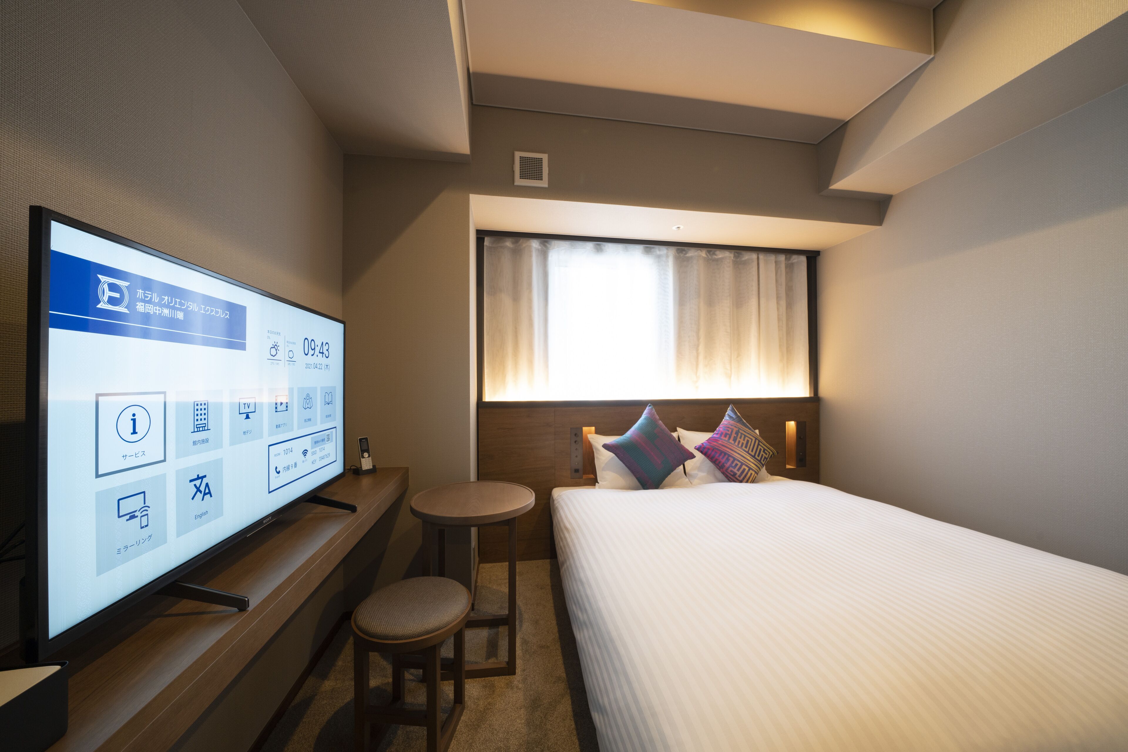 Habitación de hotel con cama, televisor sobre un soporte, una mesita pequeña y ventana con cortinas.
