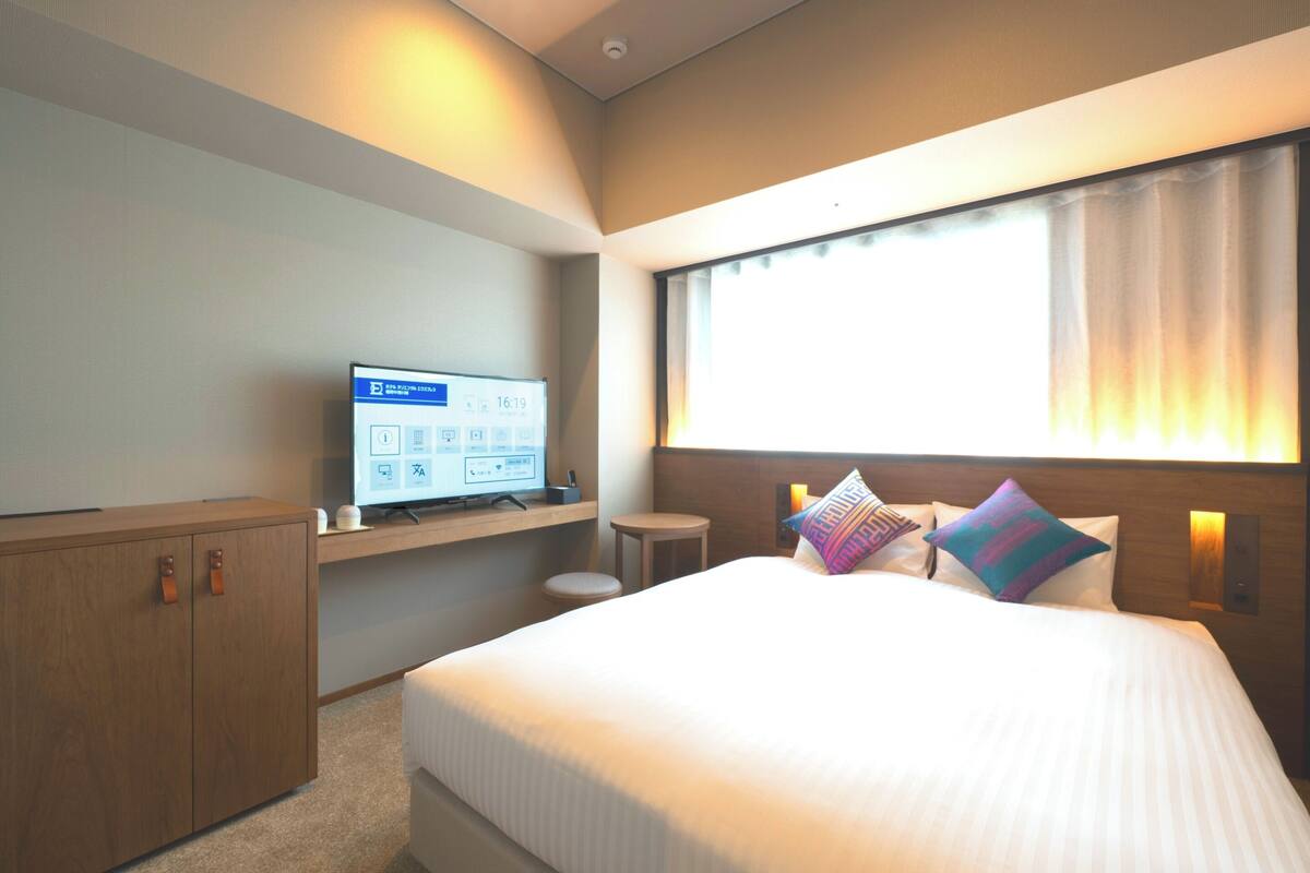 A hotel room with a bed, a desk with a computer, a wardrobe, and a window with curtains.