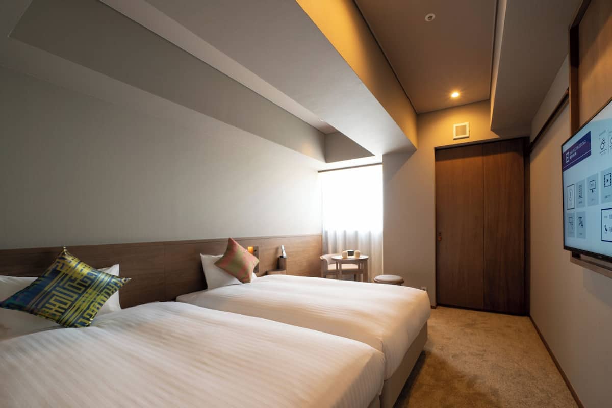 A hotel room with two beds, a small table, a chair, and a door.