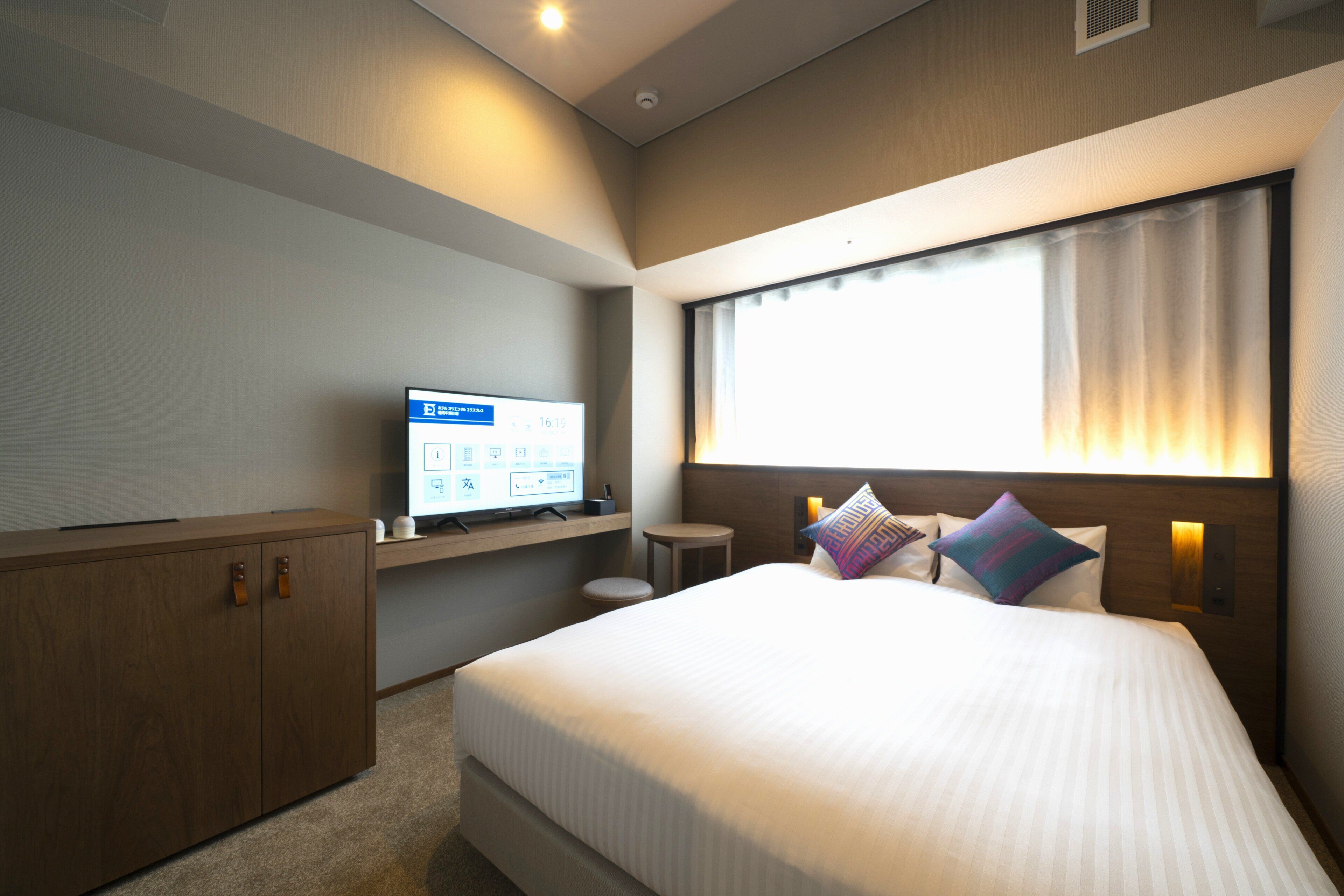Une chambre d’hôtel avec un grand lit, un téléviseur posé sur un meuble, une petite table ronde et une fenêtre avec des rideaux.