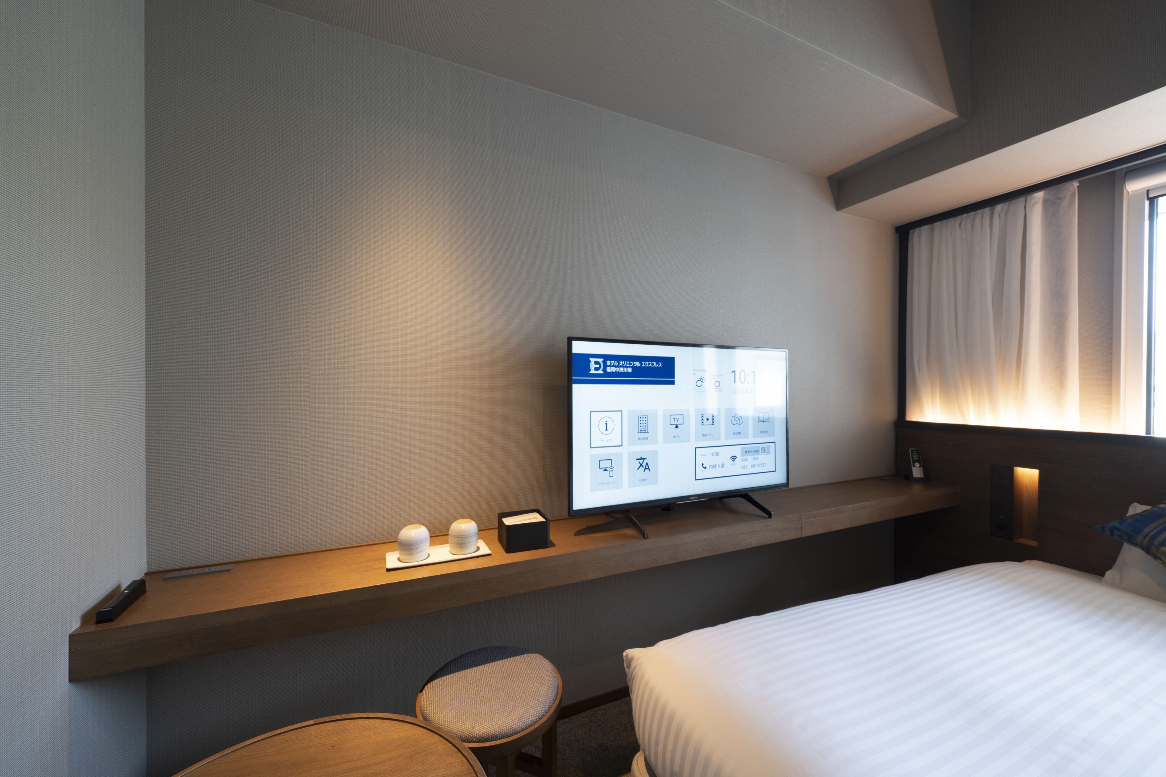 Une chambre d’hôtel comprenant un lit, un bureau avec un écran d’ordinateur affichant des informations sur la chambre, une chaise et une table de chevet avec un téléphone et une lampe.