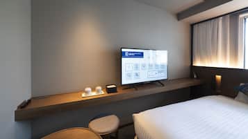 A hotel room with a bed, a desk with a computer monitor displaying room information, a chair, and a bedside table with a phone and a lamp.