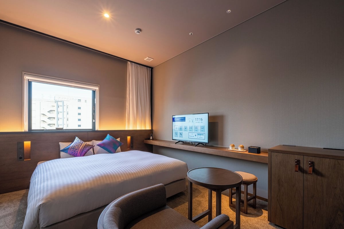 A modern hotel room with a large bed, a desk, a chair, a TV, and a window with curtains.