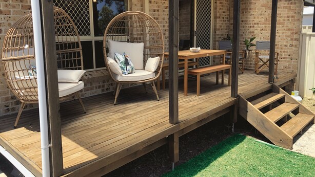 Terraza de madera con sillas de mimbre y una mesa, un muro de ladrillo y un área cubierta con un banco.
