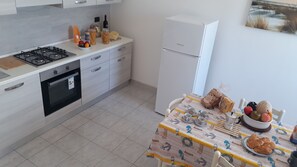 Een moderne keuken met een gasfornuis, oven en een koelkast. Er staat een eettafel met een tafelkleed en verschillende etenswaren.