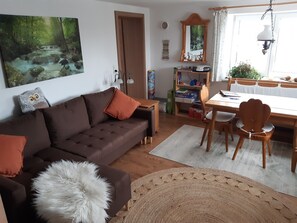 A cozy living room with a brown sofa, a white fluffy ottoman, a dining table with chairs, a bookshelf, and a large painting of a forest stream.