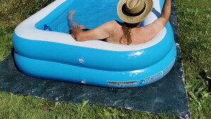 A person relaxing in a blue inflatable pool on a grassy area with a scenic view of a rural landscape and a wooden structure in the background.