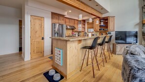 A modern kitchen with wooden cabinets, a central island with bar stools, a built-in oven, and a large TV.