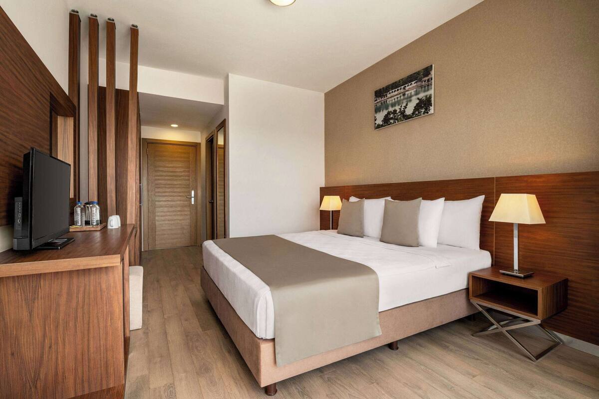 Une chambre d’hôtel avec un lit, des lampes de chevet, une télévision et un bureau en bois sur lequel se trouvent une télévision et des bouteilles d’eau.