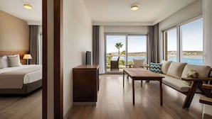 Ein Hotelzimmer mit einem Bett, einem Nachttisch aus Holz, einer Couch, einem Esstisch und Blick auf das Meer.