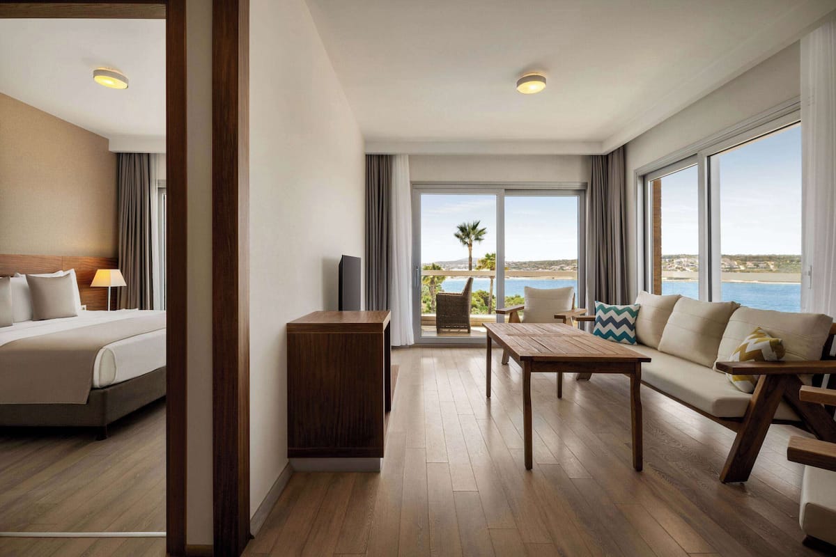 Une chambre d’hôtel avec un lit, une table de chevet en bois, un canapé, une table à manger et une vue sur l’océan.