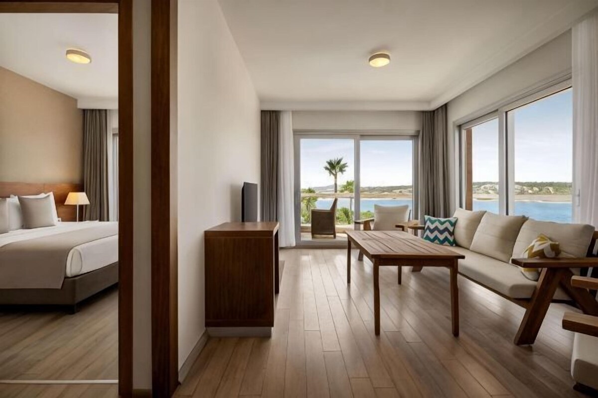 A hotel room with a bed, a sofa, a table, and a TV. There is a balcony with a view of the sea.