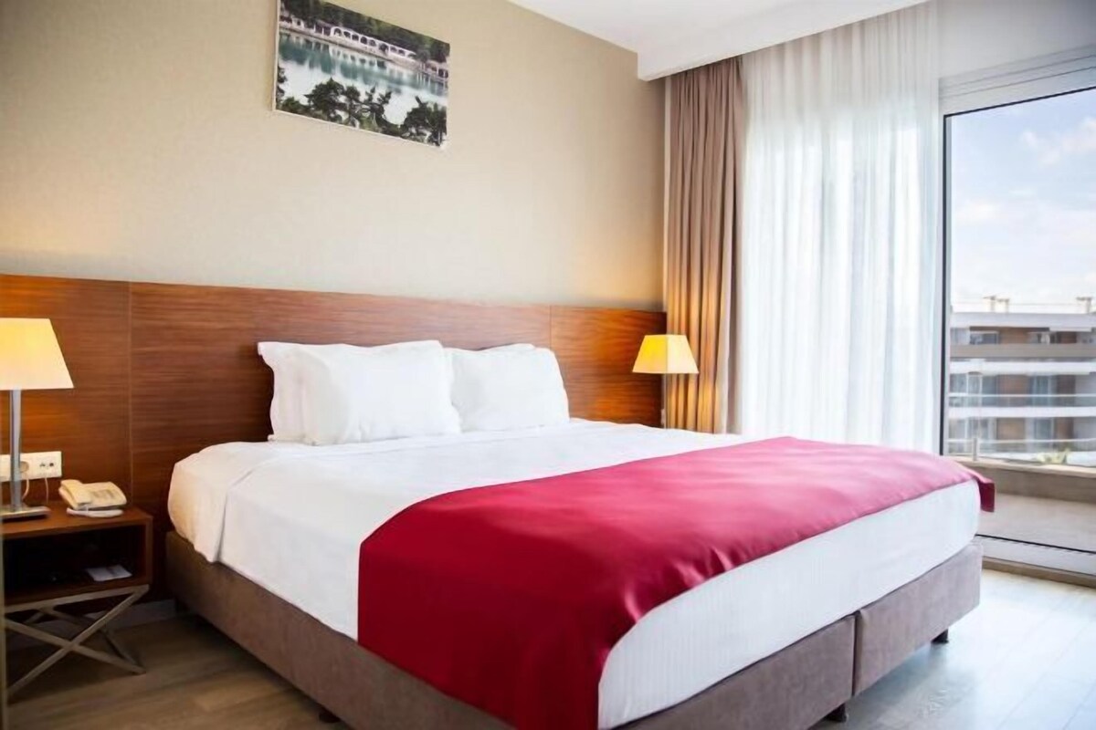 A hotel room with a large bed, two bedside lamps, a nightstand with a phone, a window with curtains, and a cityscape view.