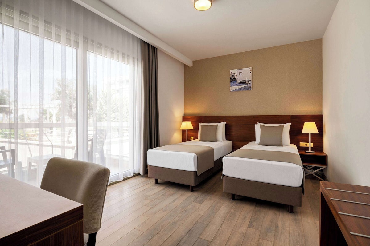 Une chambre d’hôtel avec deux lits, un bureau et une chaise. Il y a une fenêtre avec des rideaux, un tableau accroché au mur et une lampe sur chaque table de chevet.