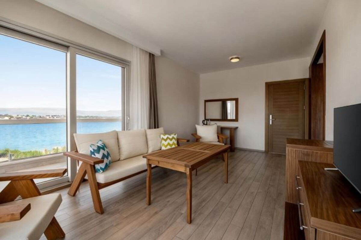 A room with a sofa, wooden coffee table, TV stand, and a large window overlooking a body of water.