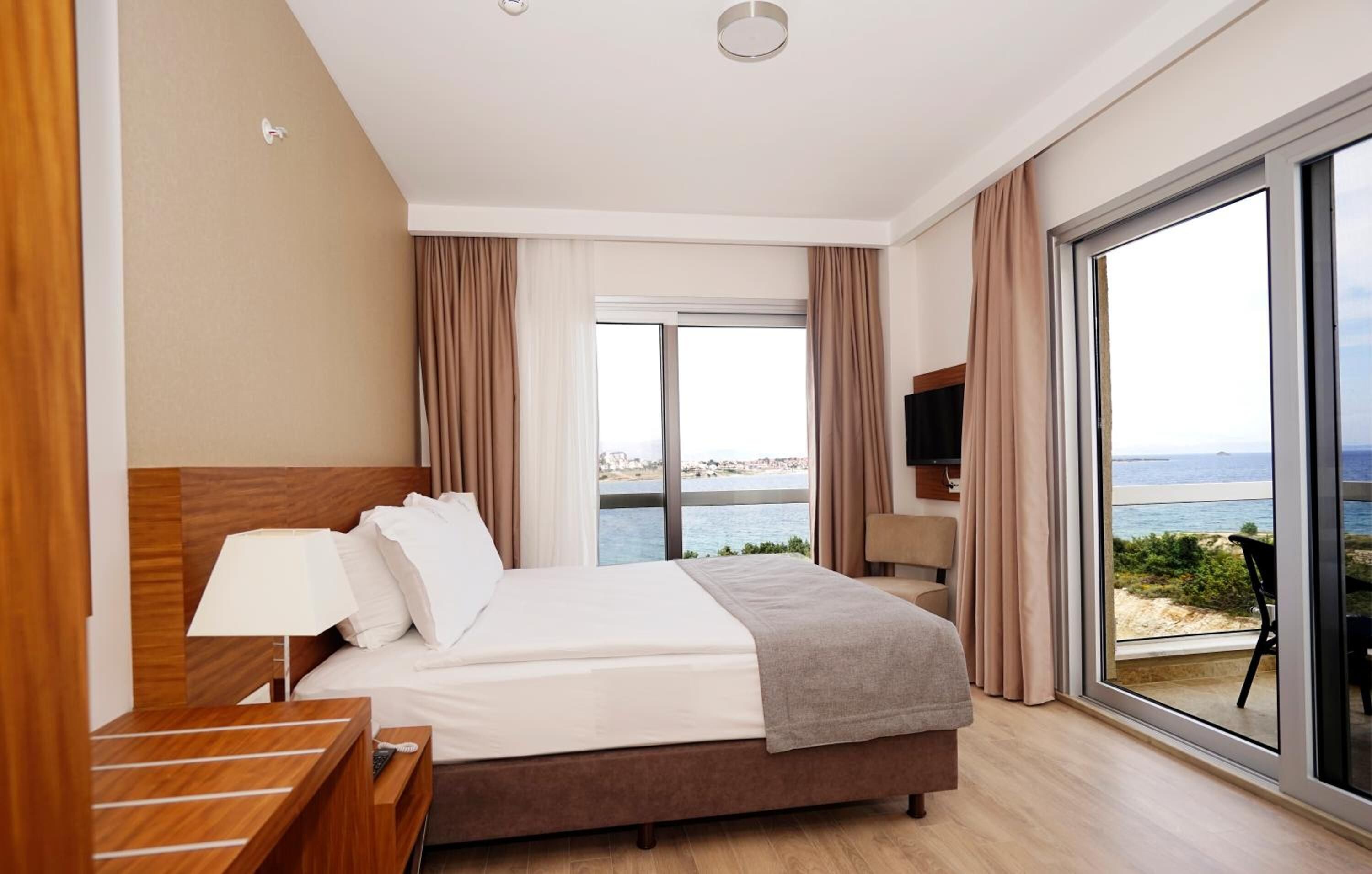 A hotel room with a large bed, a nightstand, a TV, and a balcony with a view of the sea.