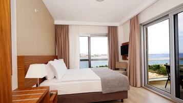 A hotel room with a large bed, a nightstand, a TV, and a balcony with a view of the sea.
