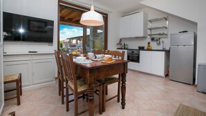 A modern kitchen with white cabinets, a dining table, and chairs. There is a TV mounted on the wall, a refrigerator, and a window with a view of the outside.