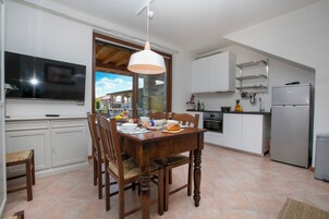 A modern kitchen with white cabinets, a dining table, and chairs. There is a TV mounted on the wall, a refrigerator, and a window with a view of the outside.