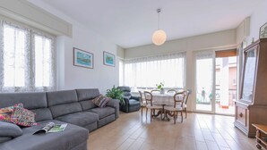 Una sala de estar con un sofá gris, una mesa de comedor con sillas y un ventanal con cortinas.