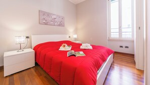 A bedroom with a red bedspread, a nightstand with a lamp, a wall art piece, and a window with blinds. - Appartamento Il Golfo: Lake View in Laveno-mombello (Laveno Mombello)