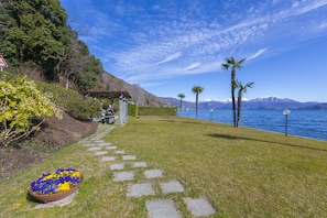 Stroll along the serene waterfront path, surrounded by lush greenery and palm trees, to a tranquil outdoor space beneath a clear blue sky.