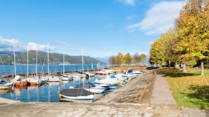 A marina with boats docked along a stone quay, a clear sky, and autumn foliage in the background. - Riva del Caolino (Ispra)