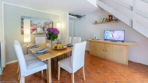 A dining area with a wooden table, white chairs, a vase with flowers, and a television on a wooden cabinet. - Riva del Caolino (Ispra)