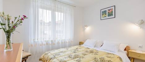 A bedroom with a bed, bedside table, lamp, and a window with sheer curtains.