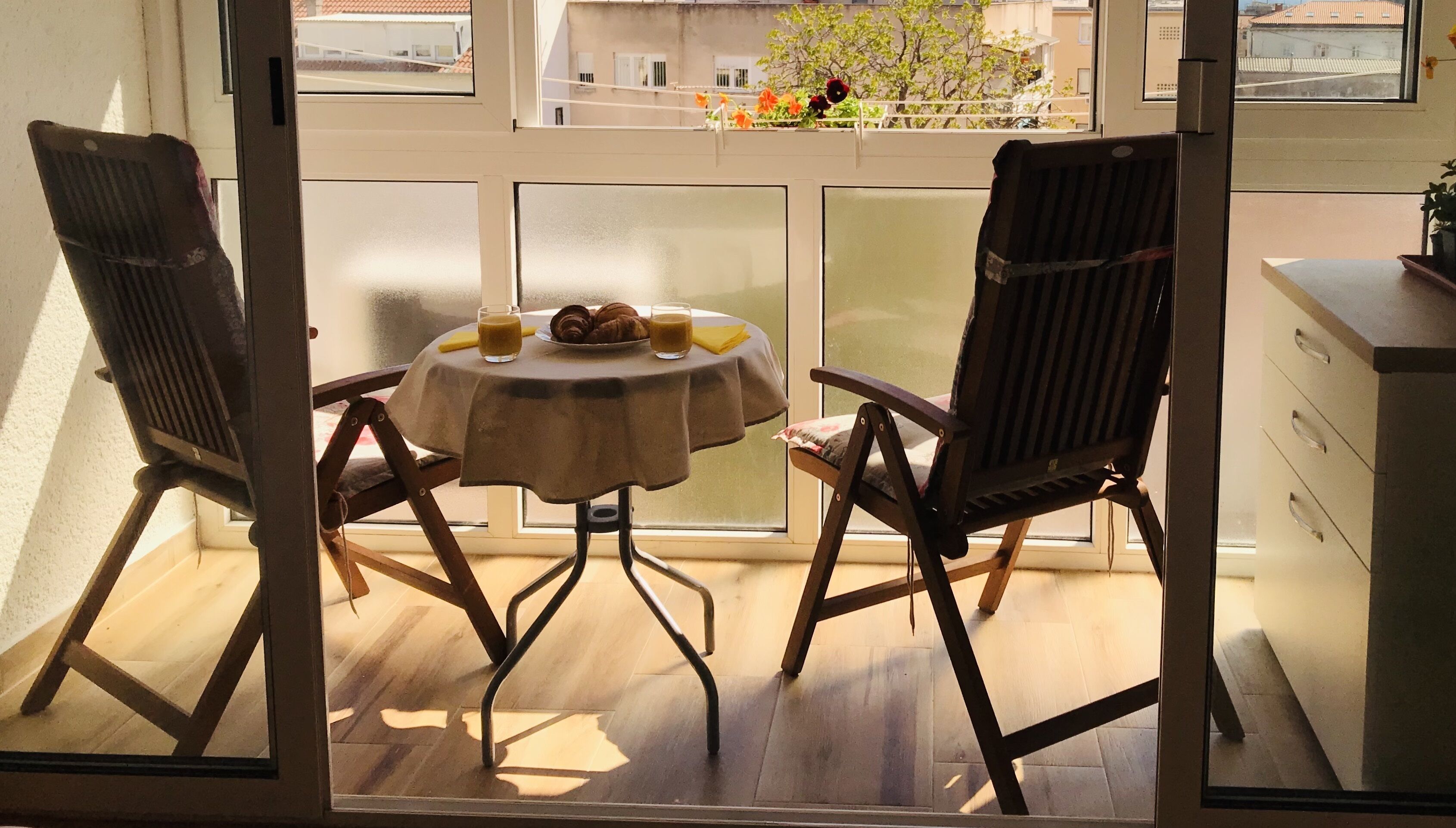 A balcony with two chairs, a table with breakfast items, and a view of buildings and trees.