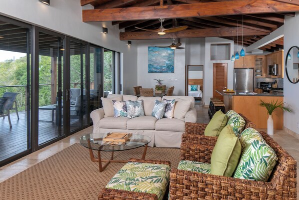 A modern living room with a sofa, ottoman, and glass coffee table. There is a kitchen area with a refrigerator and microwave. The room has large windows and a ceiling fan. - Eureka Villa -Stunning Views in Paradise 2BR/2.5BA (Rendezvous Bay)