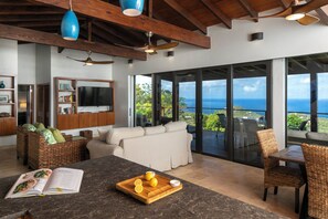 A modern living room with a large window, a dining area, and a kitchen counter with lemons. - Eureka Villa -Stunning Views in Paradise 2BR/2.5BA (Rendezvous Bay)