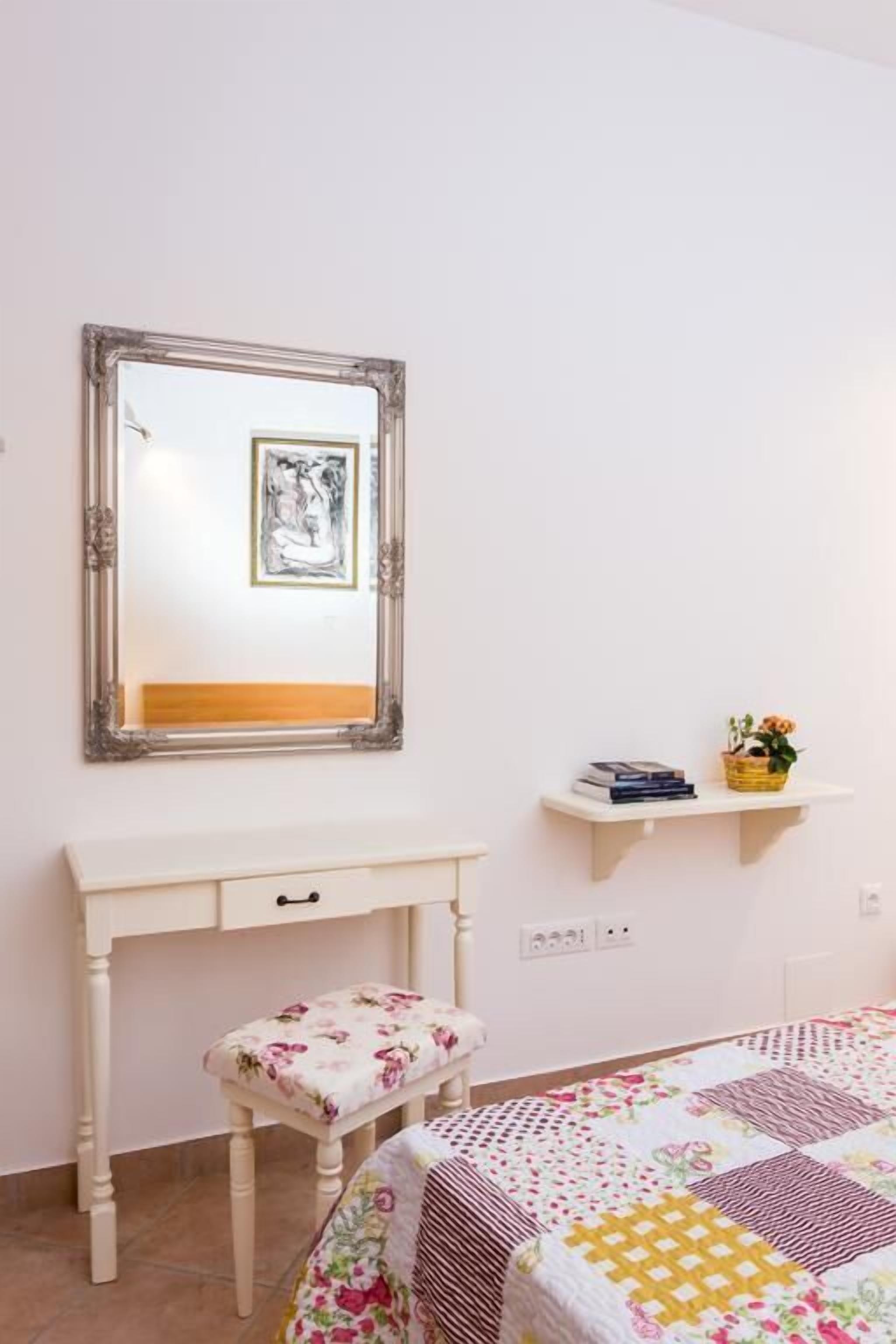 A bedroom with a bed, a bedside table, a mirror, and a small shelf with books and a plant.