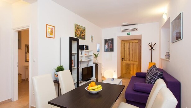 Un coin repas avec une table, des chaises et un saladier de fruits. On aperçoit un canapé violet et une porte en bois à l’arrière-plan.