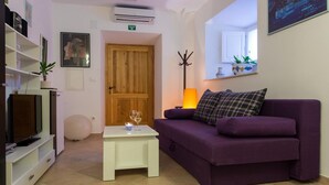 Un salon avec un canapé violet, une petite table blanche sur laquelle sont posés des verres, une porte en bois et un téléviseur placé sur un meuble blanc.