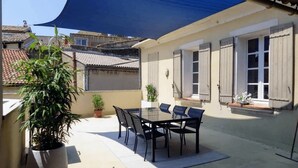 Un patio al aire libre con una mesa y sillas, rodeado de plantas en macetas y un toldo azul.