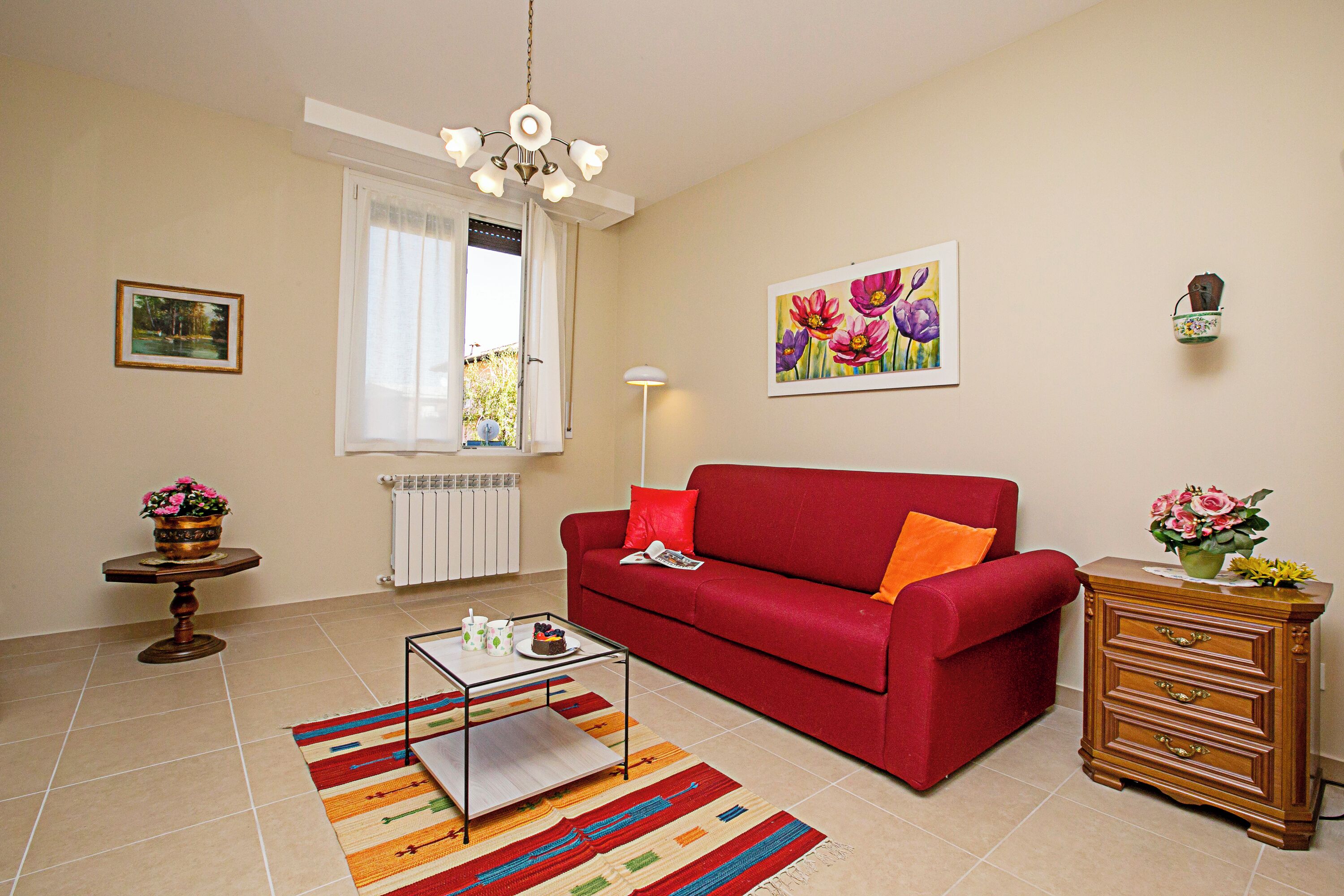 Ein Wohnzimmer mit einem roten Sofa, einem Couchtisch, einem gemusterten Teppich, einem Holznachttisch mit Blumen und einem Gemälde an der Wand.
