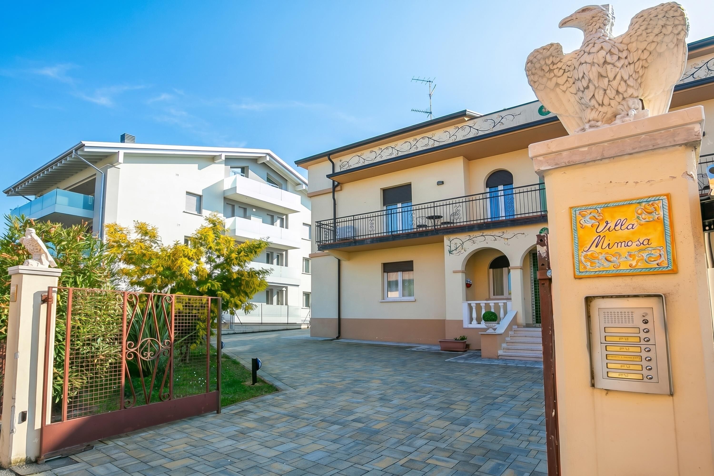 Une courClose agrémentée d’une villa appelée « Villa Mimosa », d’une statue d’aigle en pierre et, au fond, d’un bâtiment moderne.