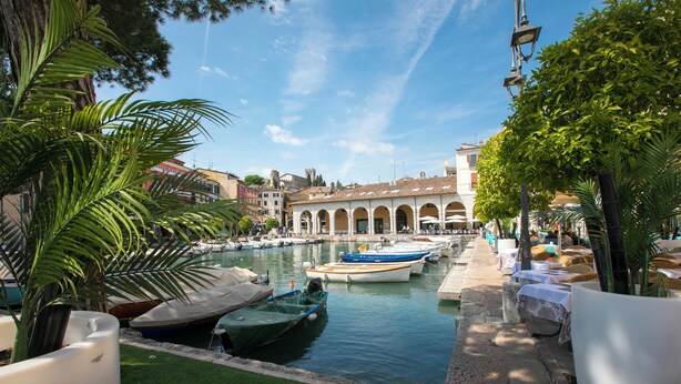 A canal with boats docked, a building with arches, and a row of outdoor seating along the water.